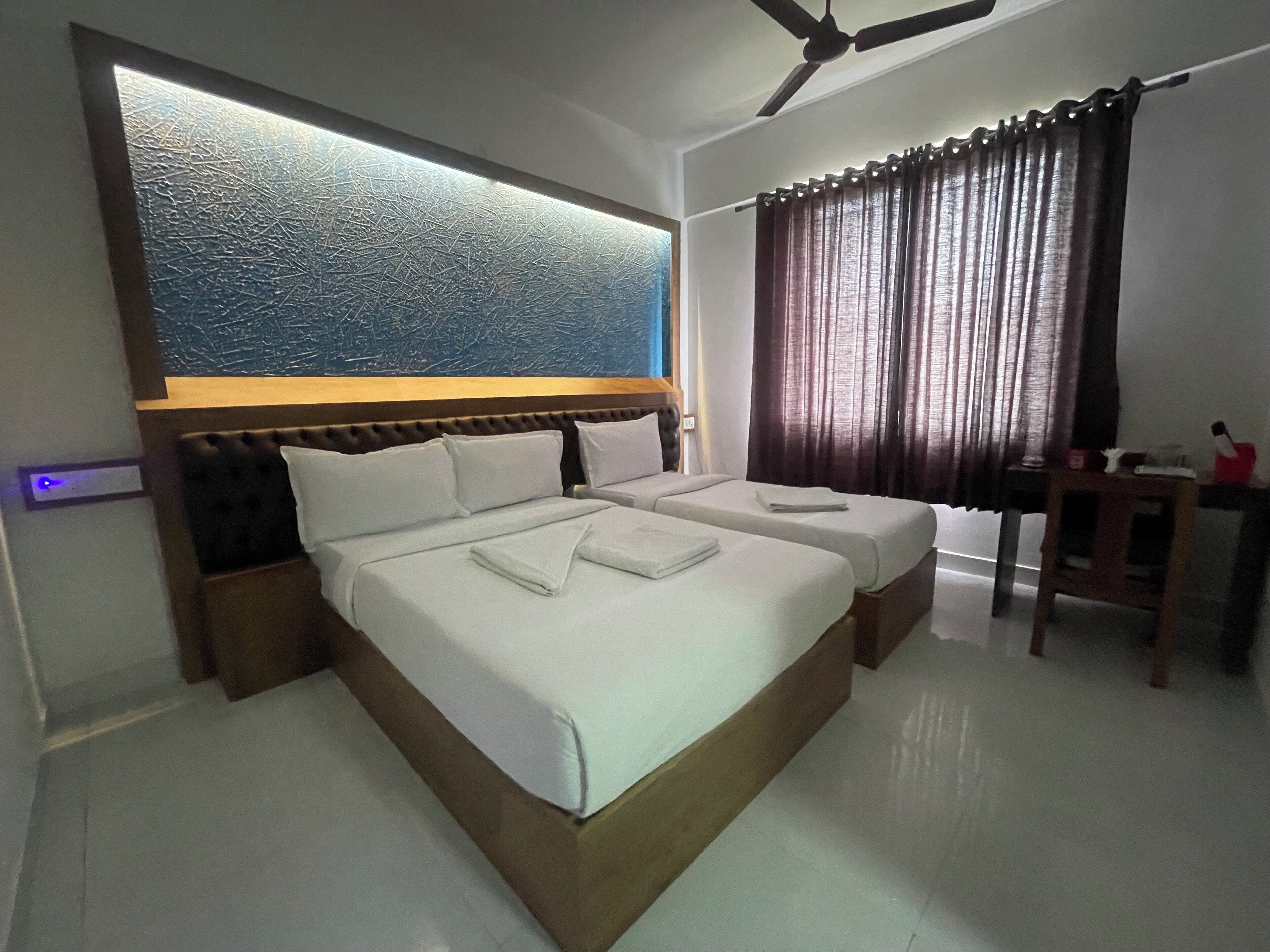 A hotel room with two beds, a ceiling fan, a nightstand, a table, and a curtain.