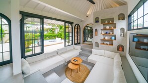 A spacious living area with a white sofa set, a small wooden table, and a ceiling fan. Large windows provide a view of the outdoor pool and greenery. - Villa Mellow (Canggu)