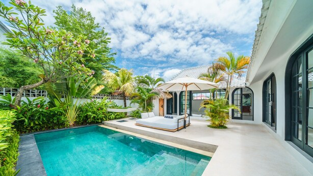 A modern outdoor pool area with a lounge chair and umbrella, surrounded by lush greenery and a building with large windows. - Villa Mellow (Canggu)