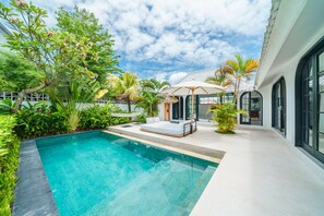 Un área de piscina exterior moderna con una silla de descanso y sombrilla, rodeada de abundante vegetación y un edificio con grandes ventanales.