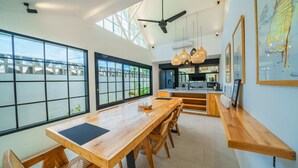 A spacious dining area with a long wooden table and chairs, large windows, and a modern kitchen in the background. - Villa Mellow (Canggu)