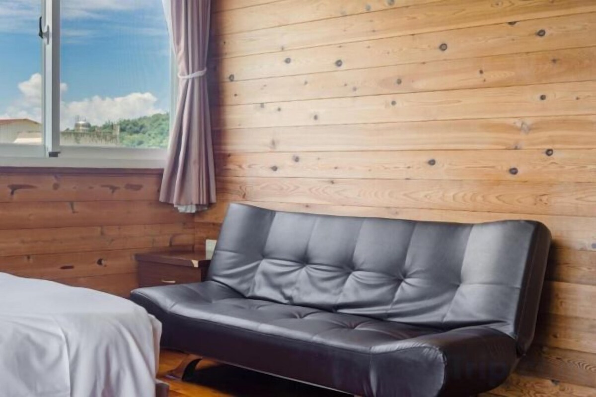 Una habitación con pared de madera, sofá de cuero negro, mesita de noche pequeña y ventana con cortinas.