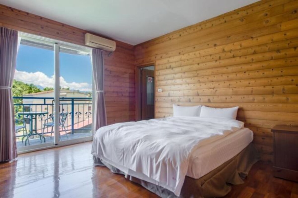 Una habitación con paneles de madera, una cama, un ventanal corredero a un balcón y una pequeña mesa con sillas afuera.