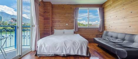 A room with a bed, a sofa, wooden walls, and a view of a body of water and buildings.