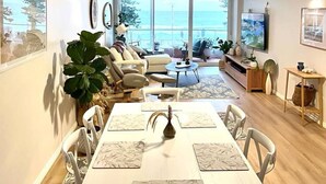 A spacious living room with a large window overlooking the ocean, a dining area with a white table and chairs, and a seating area with a sofa and armchairs. - Stunning, uninterrupted views of beach & surf (Manly)