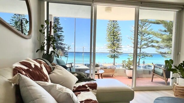 A living room with a sofa, a dining table, and a view of the ocean through large glass doors. - Stunning, uninterrupted views of beach & surf (Manly)