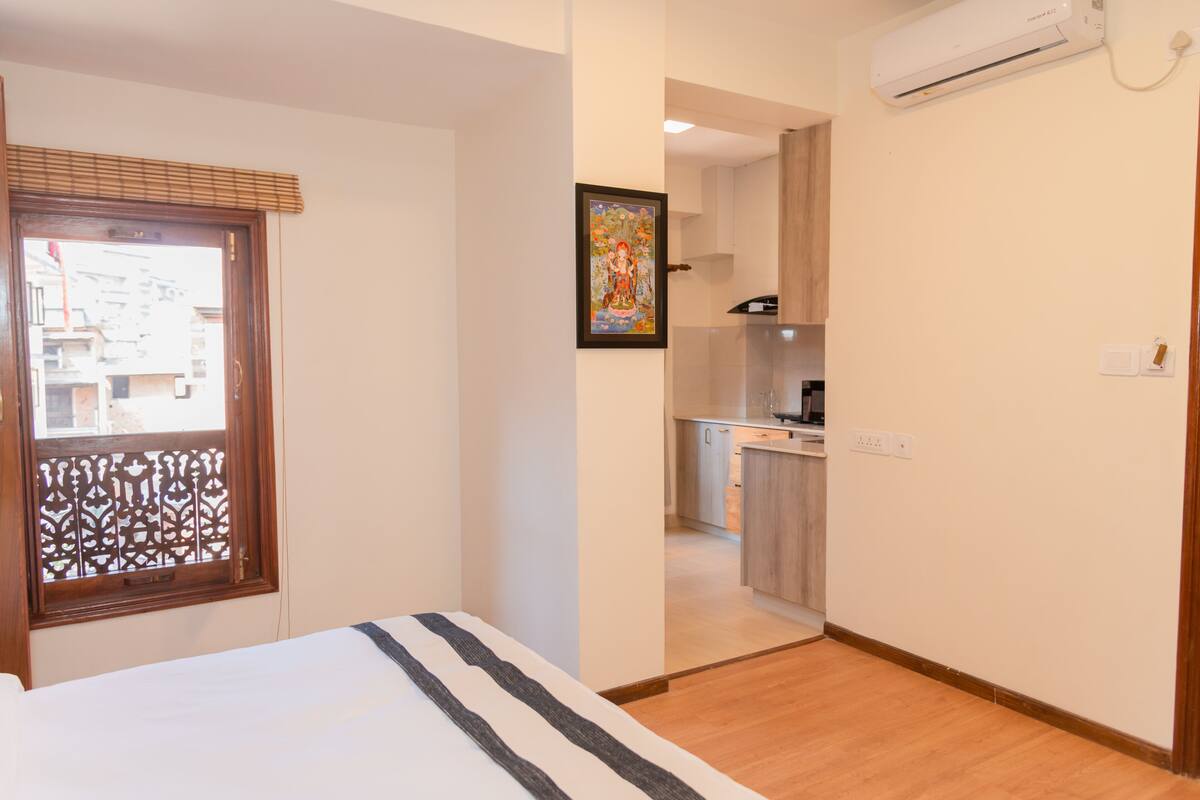 Un dormitorio con cama, una ventana con un panel decorativo, una zona de cocina visible a través de una puerta abierta y un equipo de aire acondicionado instalado en la pared.