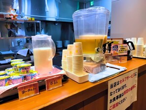 Una estación de bebidas con dispensador de leche, una jarra grande de un líquido amarillo y pilas de vasos desechables.