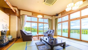 A traditional Japanese-style room with tatami mats, a low table, two chairs, and large windows offering a view of greenery. - Takasago onsen (Asahikawa)