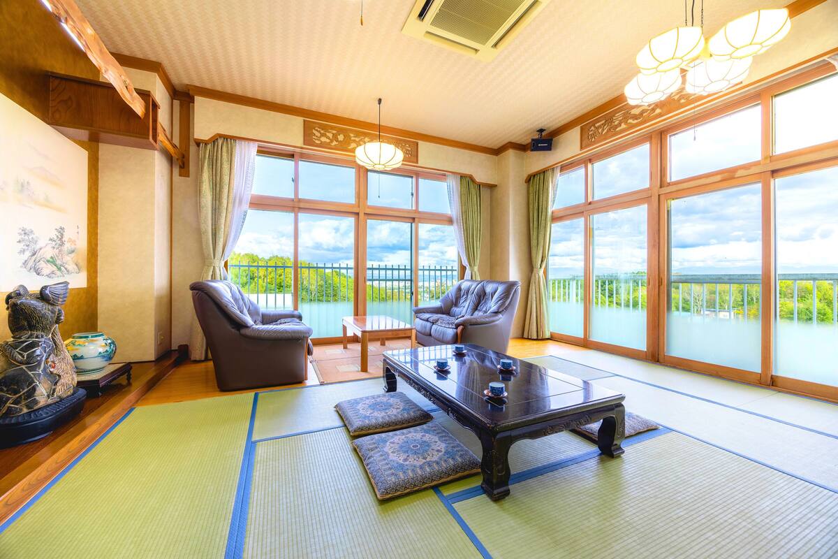 A traditional Japanese-style room with tatami mats, a low table, two chairs, and large windows offering a view of greenery.