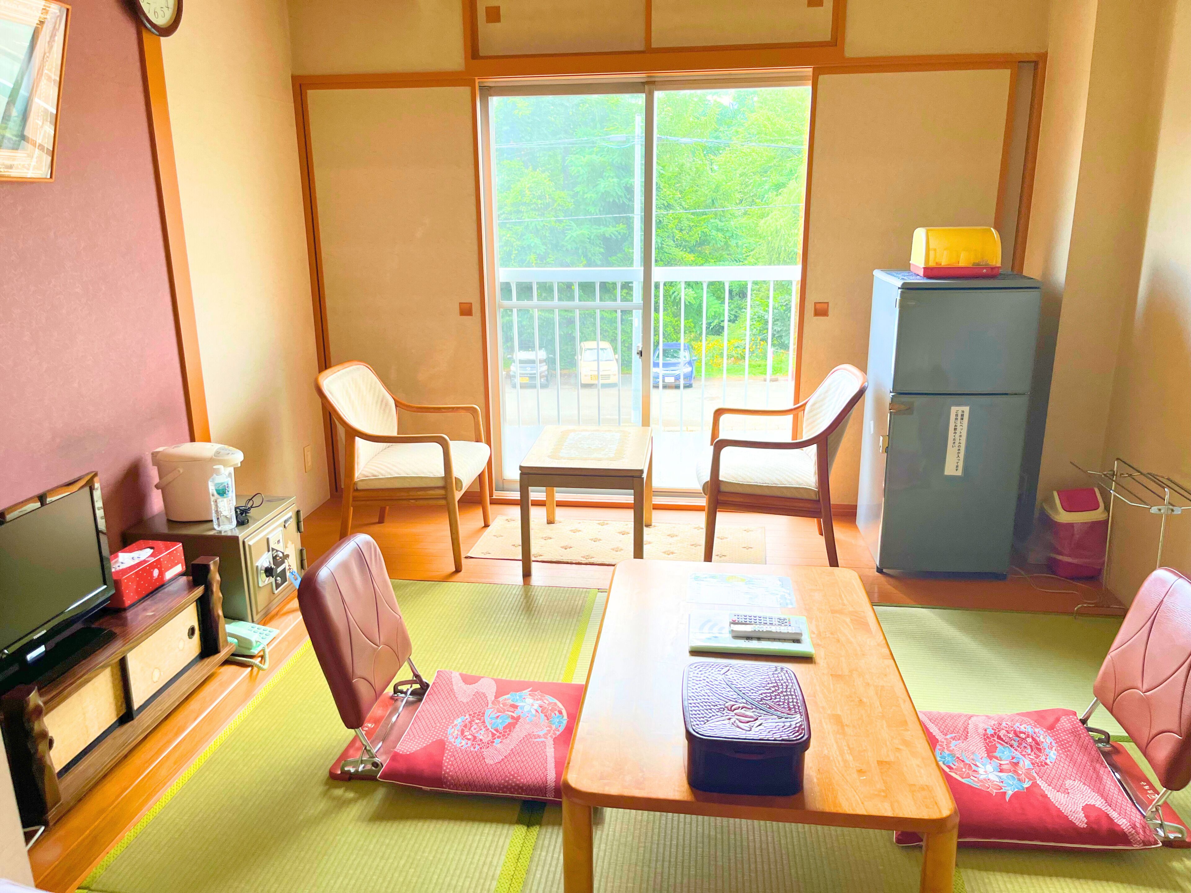 Une pièce de style japonais traditionnel, avec un sol recouvert de tatamis, une table basse, deux chaises, un téléviseur, un réfrigérateur et une porte coulissante donnant sur un balcon.