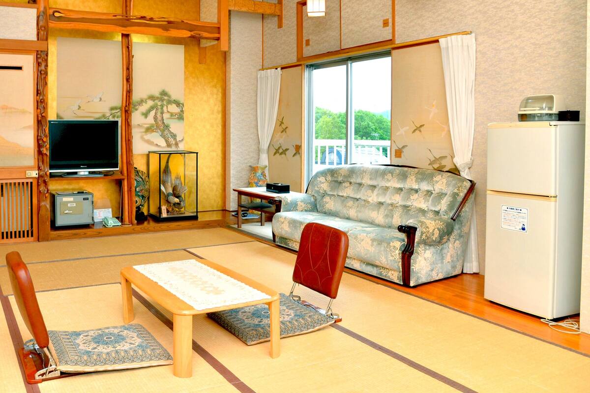 A traditional Japanese-style living room with tatami flooring, a low table, a sofa, a television, and a refrigerator.