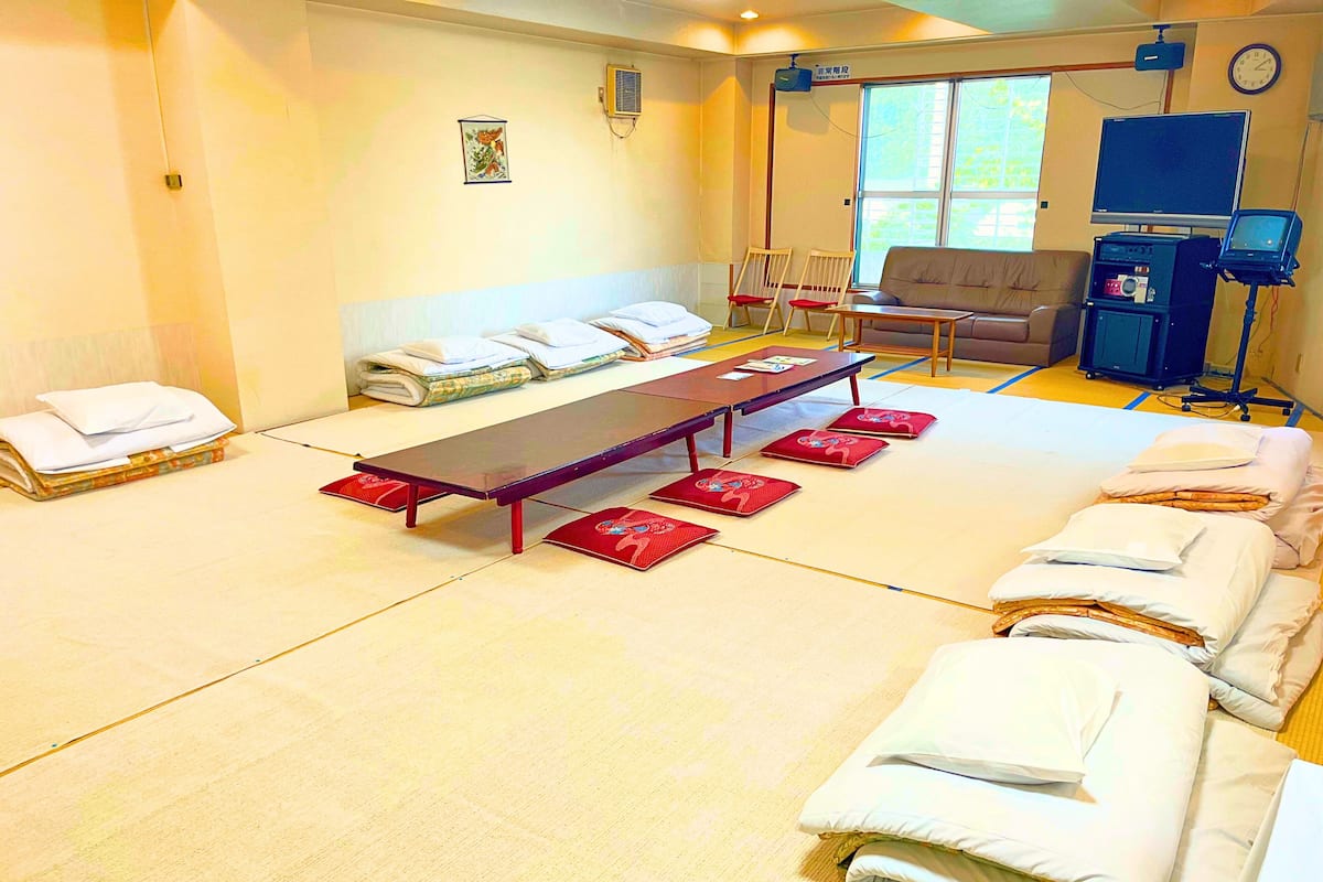 A tatami mat room with a long table, chairs, and neatly folded bedding.