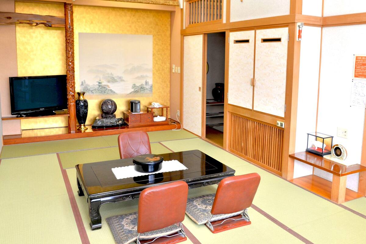 A traditional Japanese room with tatami flooring, a low table with a black tabletop, two red chairs, a television, a painting, and a cabinet with a small safe.