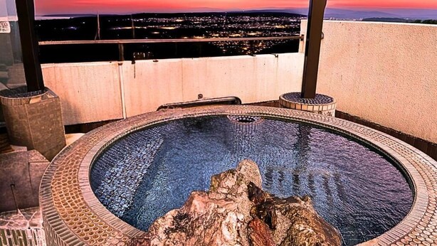A hot tub with a rocky centerpiece, surrounded by a tiled border, set against a sunset cityscape view. - Takasago onsen (Asahikawa)