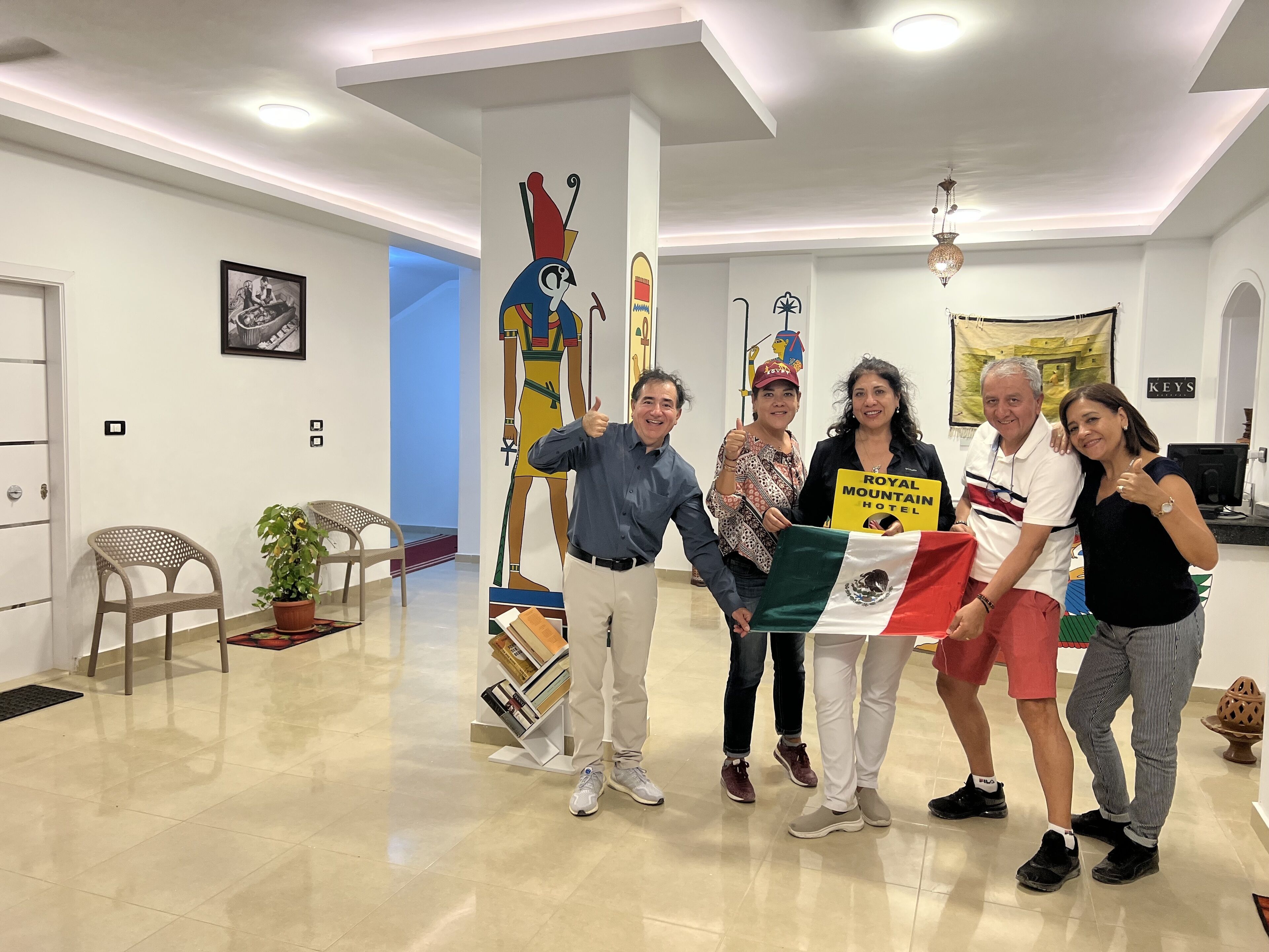 Un grupo de personas en un vestíbulo bien iluminado con diseño moderno, que incluye un pilar grande con obras de arte, una zona de estar con sillas y un cartel del Royal Mountain Hotel.