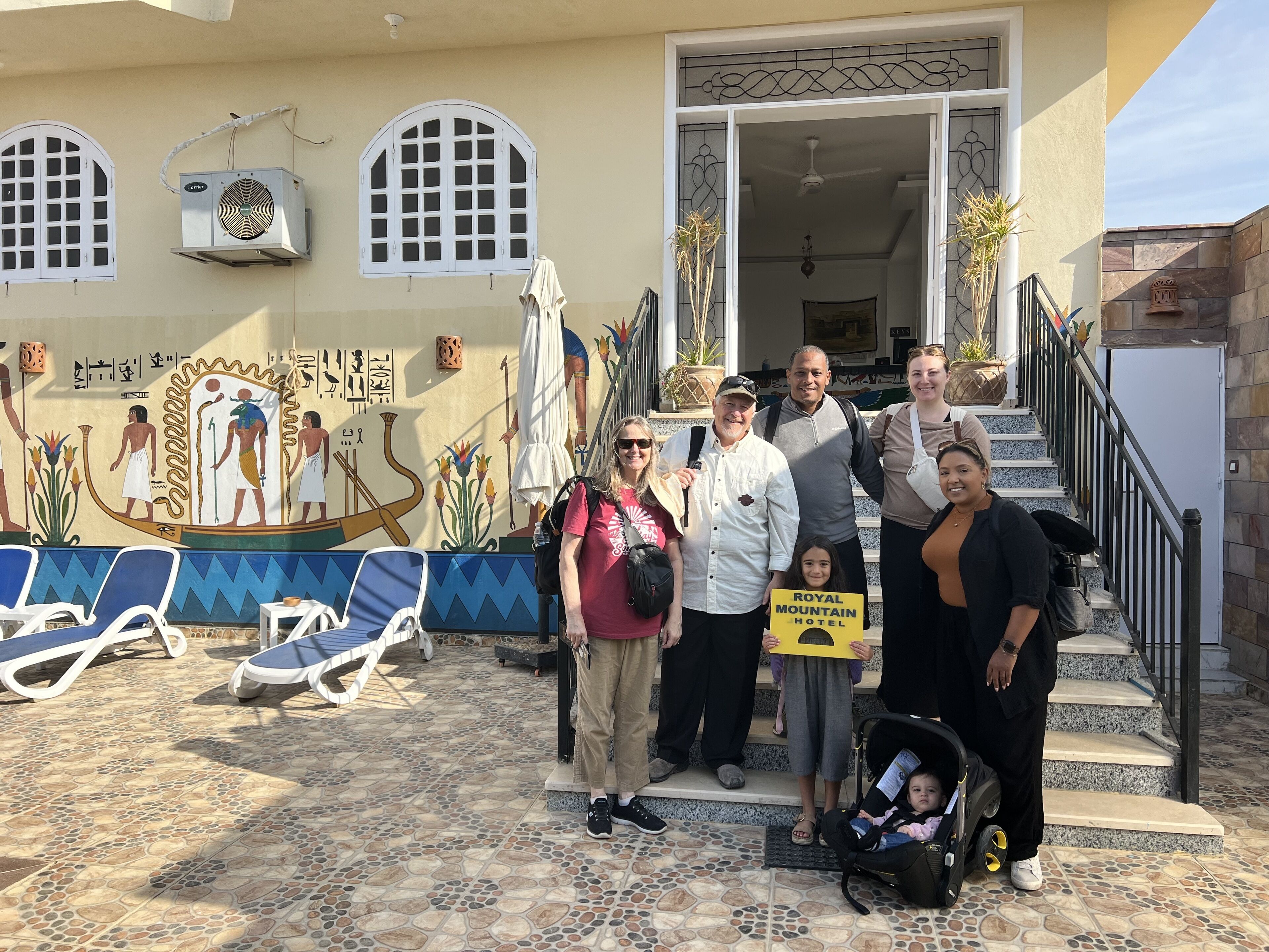 A group of people outside a building with a mural, a sign for the Royal Mountain Hotel, and a tiled patio with lounge chairs.