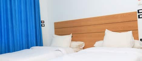 Una cama doble con ropa de cama blanca, cabecera de madera, cortina azul y lámpara fijada a la pared.