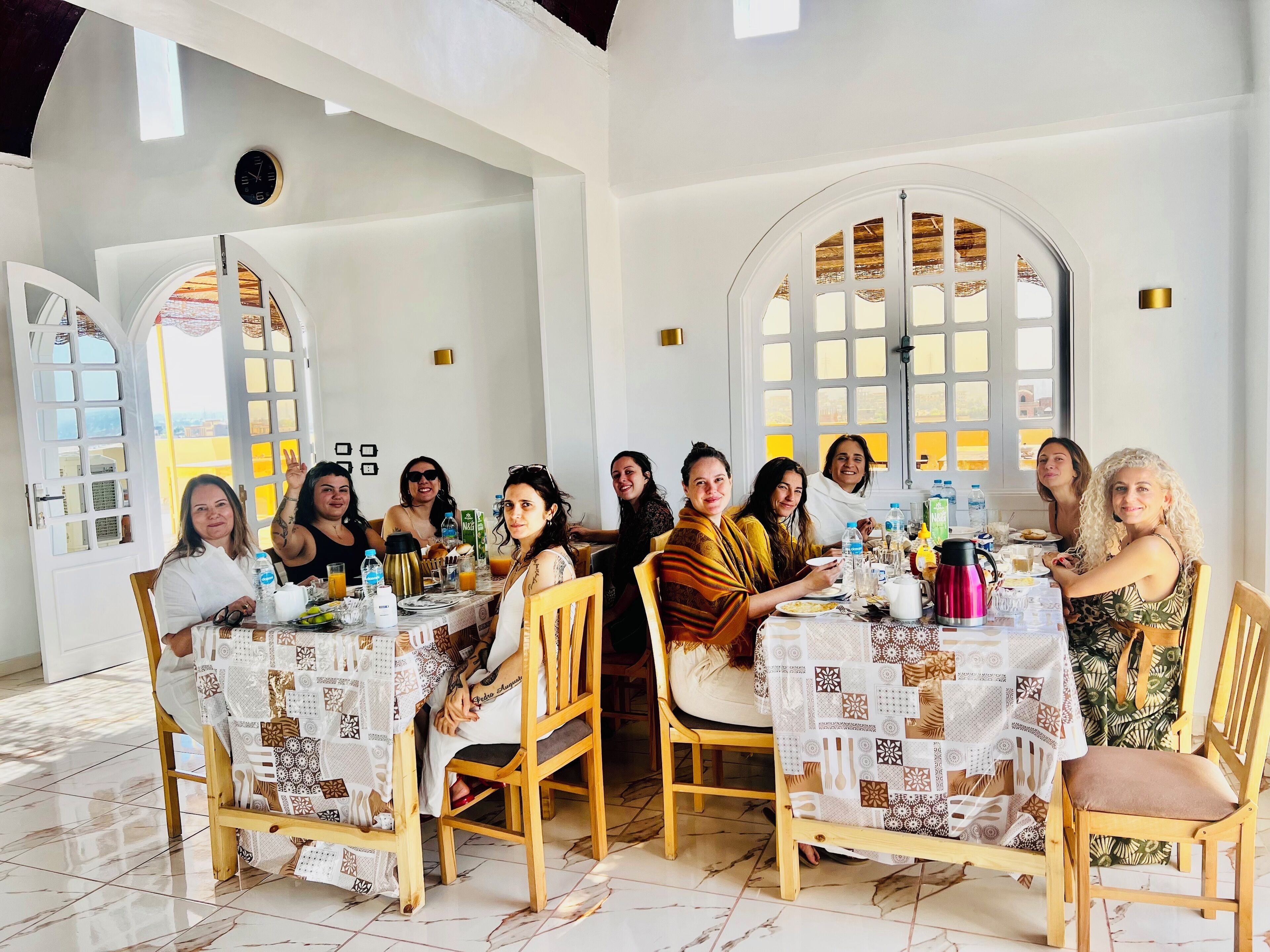 A group of people dining at a table with patterned tablecloths, wooden chairs, and a tiled floor.
