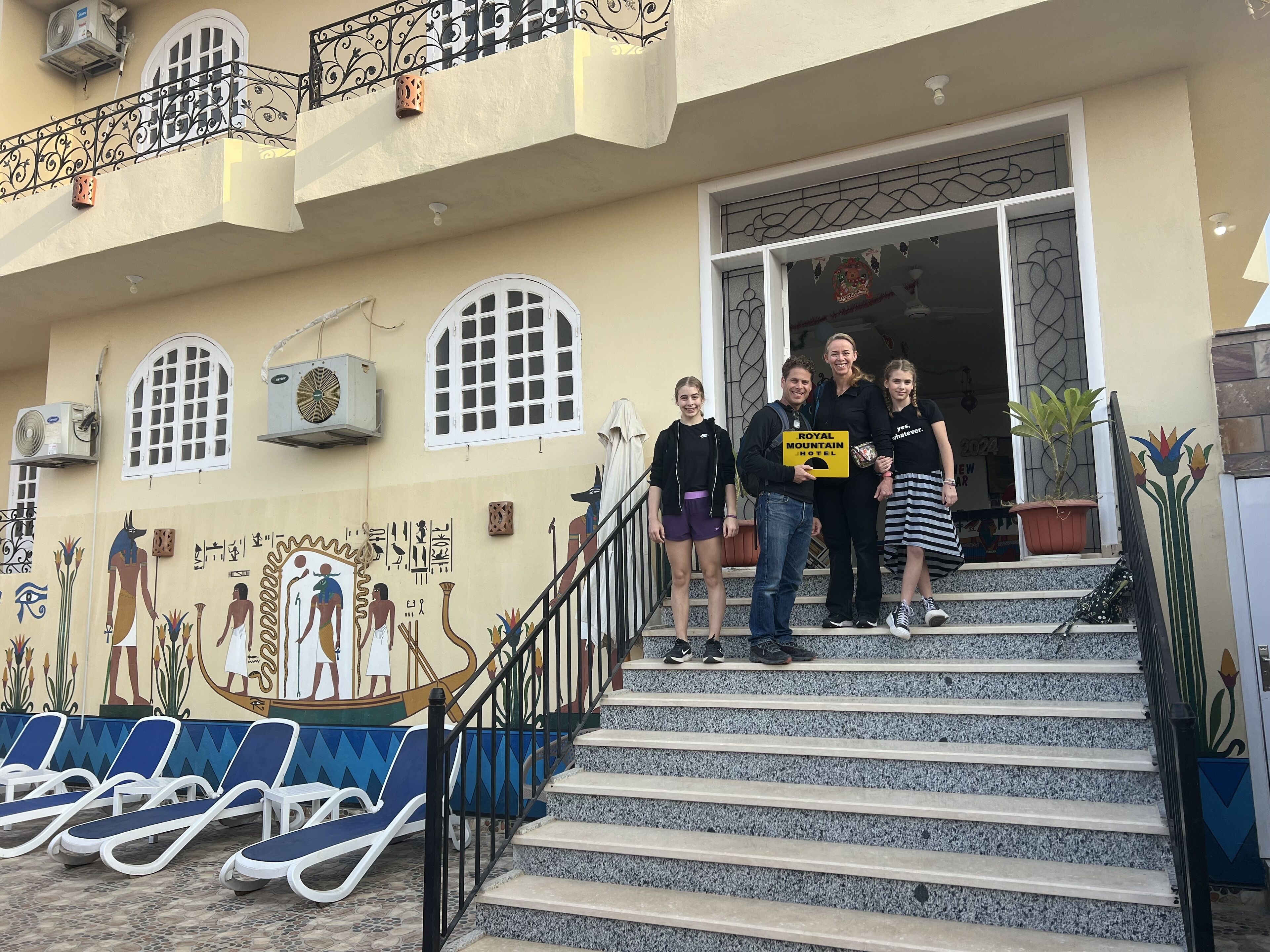 L’entrée d’un hôtel, avec une enseigne indiquant « Royal Mountain Hotel », un escalier et une fresque représentant des personnages égyptiens sur le mur.