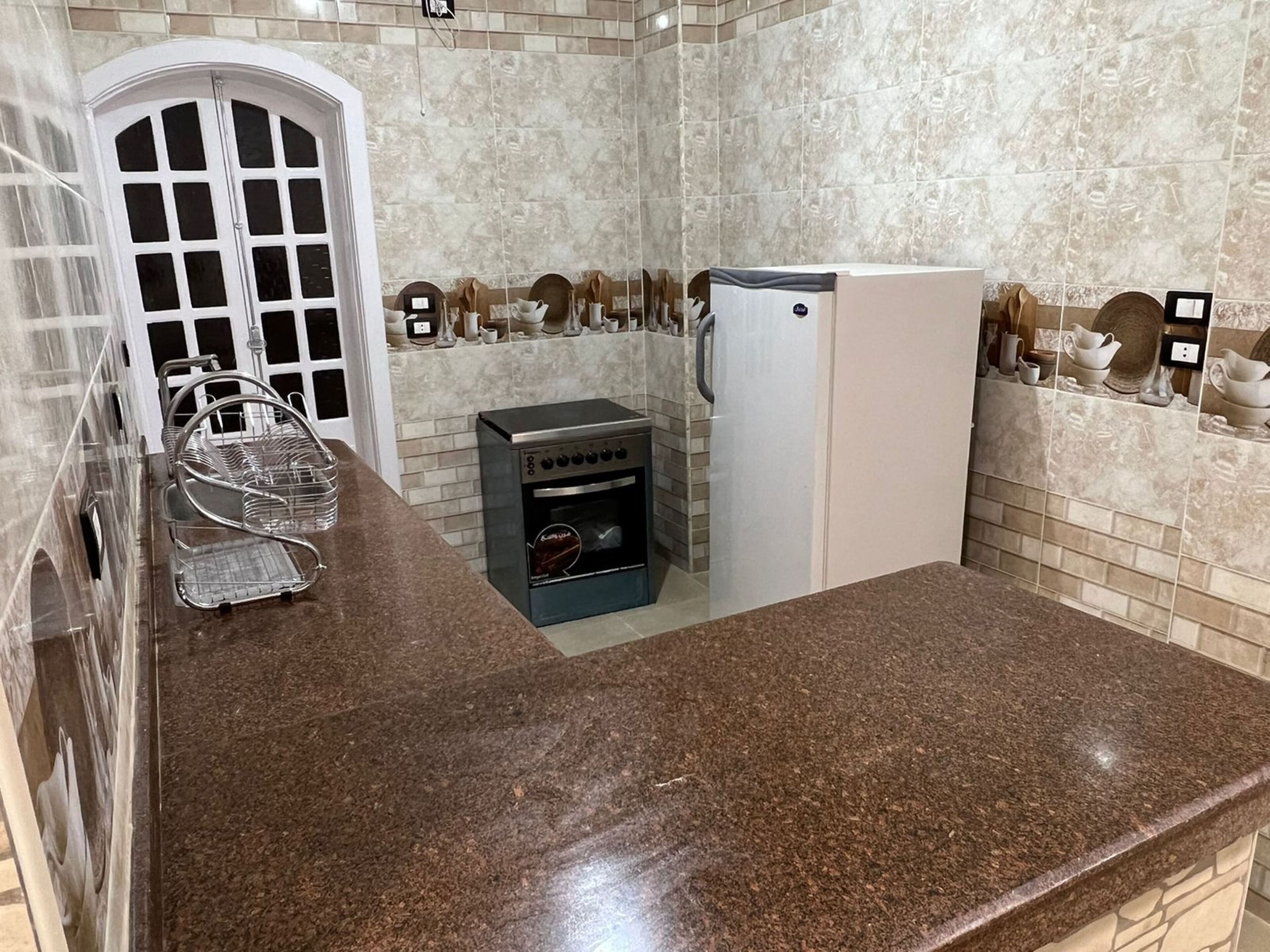A compact kitchen with a granite countertop, a built-in oven, a refrigerator, and a white door.