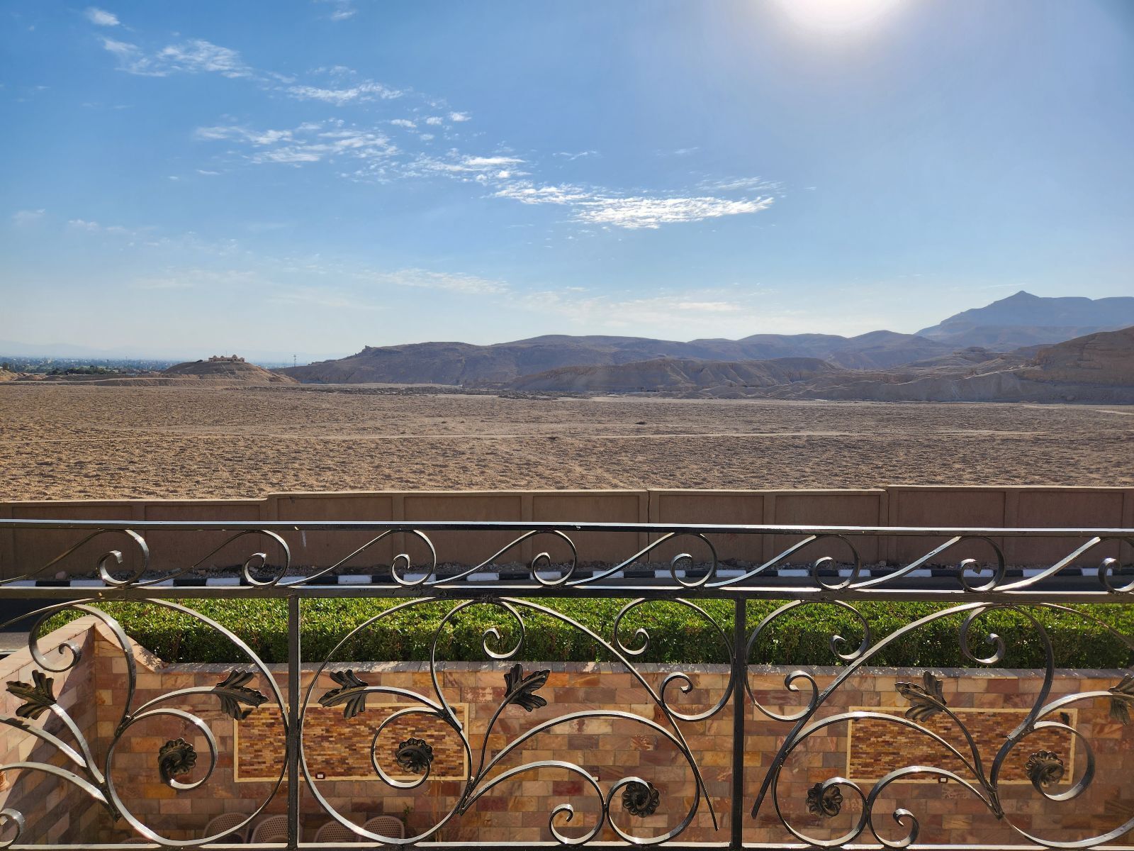 A balcony with a metal railing and decorative elements, overlooking a desert landscape with distant mountains and a clear sky.