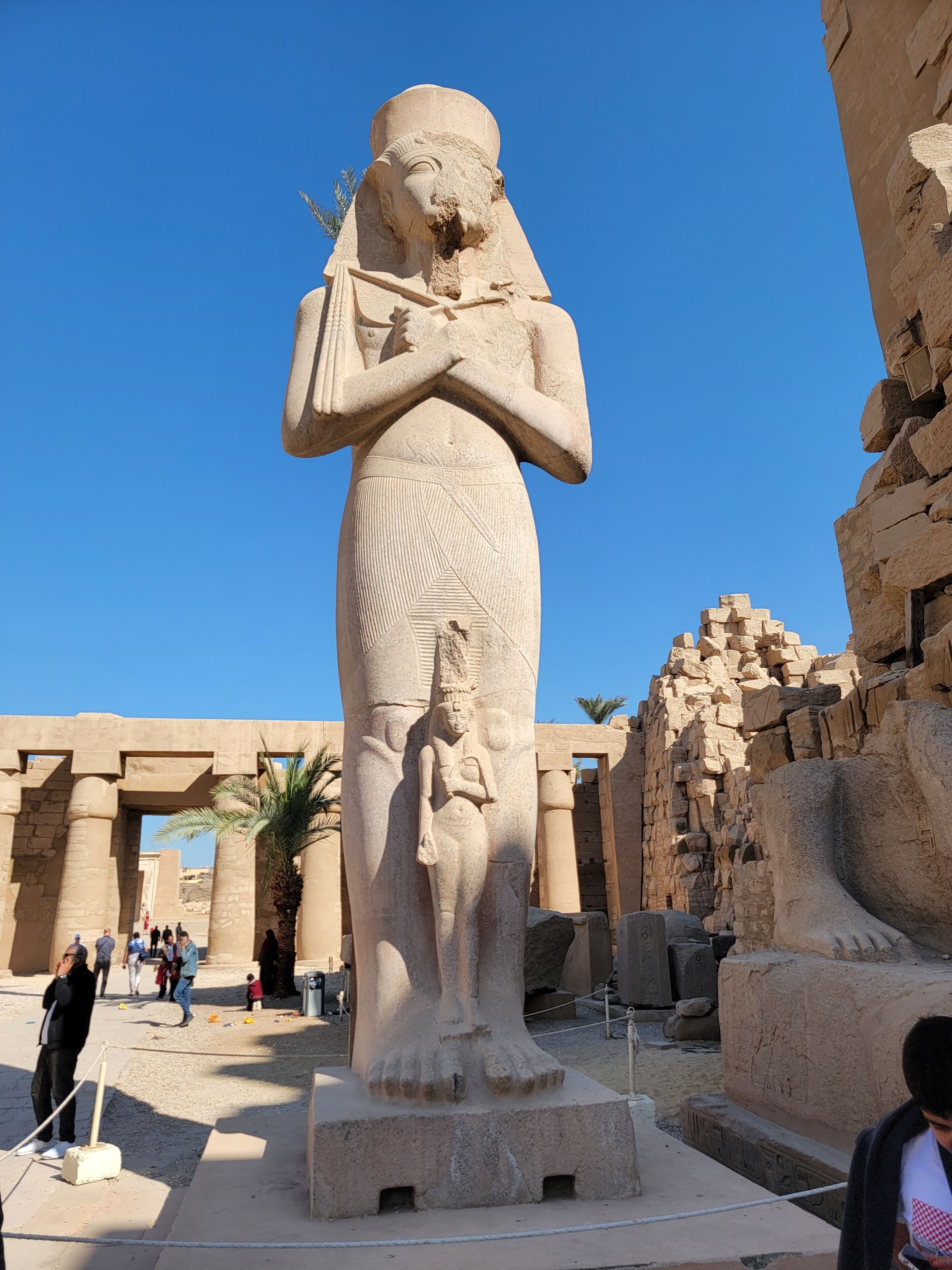 A large stone statue of a pharaoh with a tall, narrow base, surrounded by ancient stone structures and palm trees.