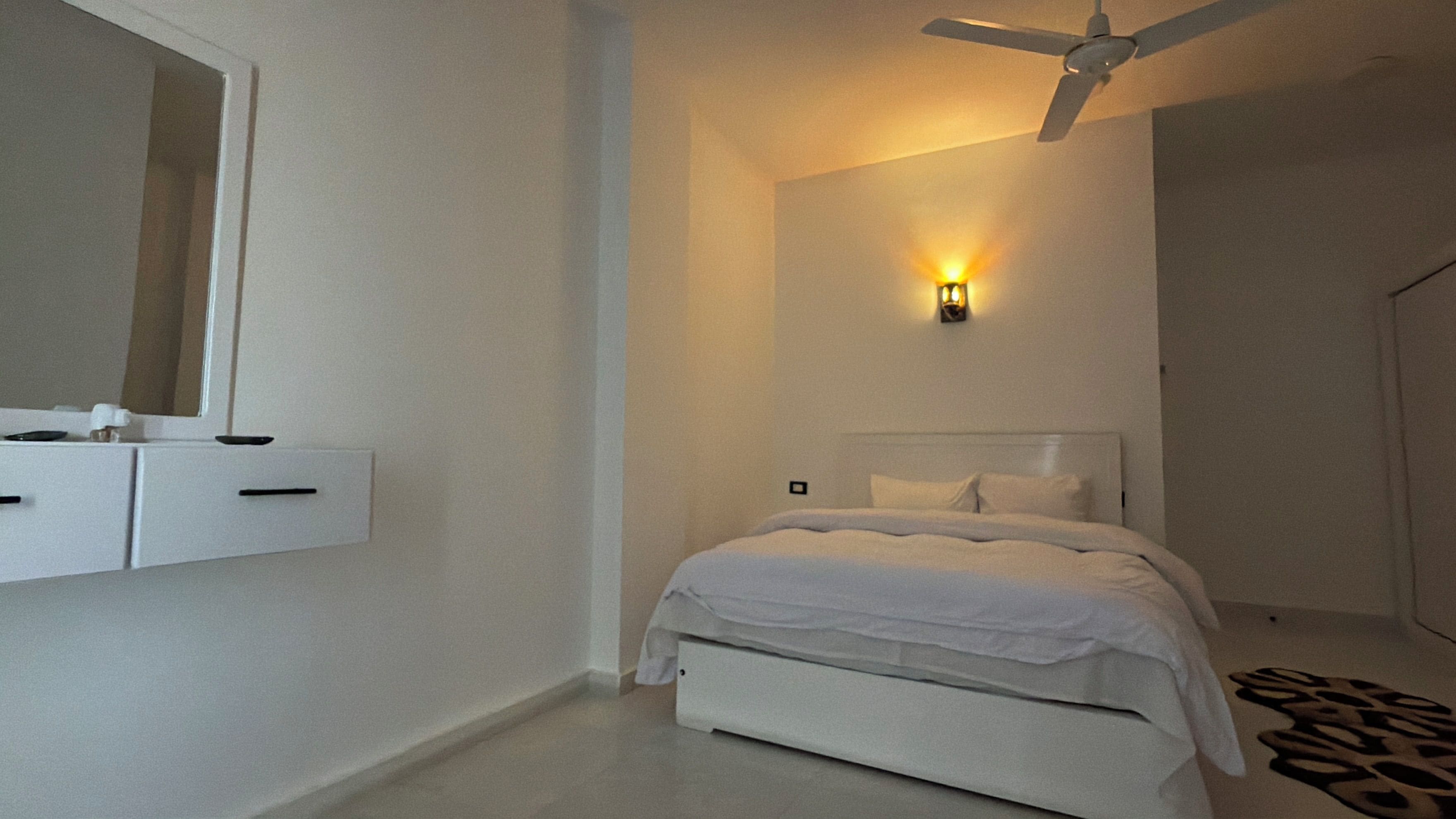 Un dormitorio con una cama, un ventilador de techo, una lámpara fijada a la pared y un aparador blanco.
