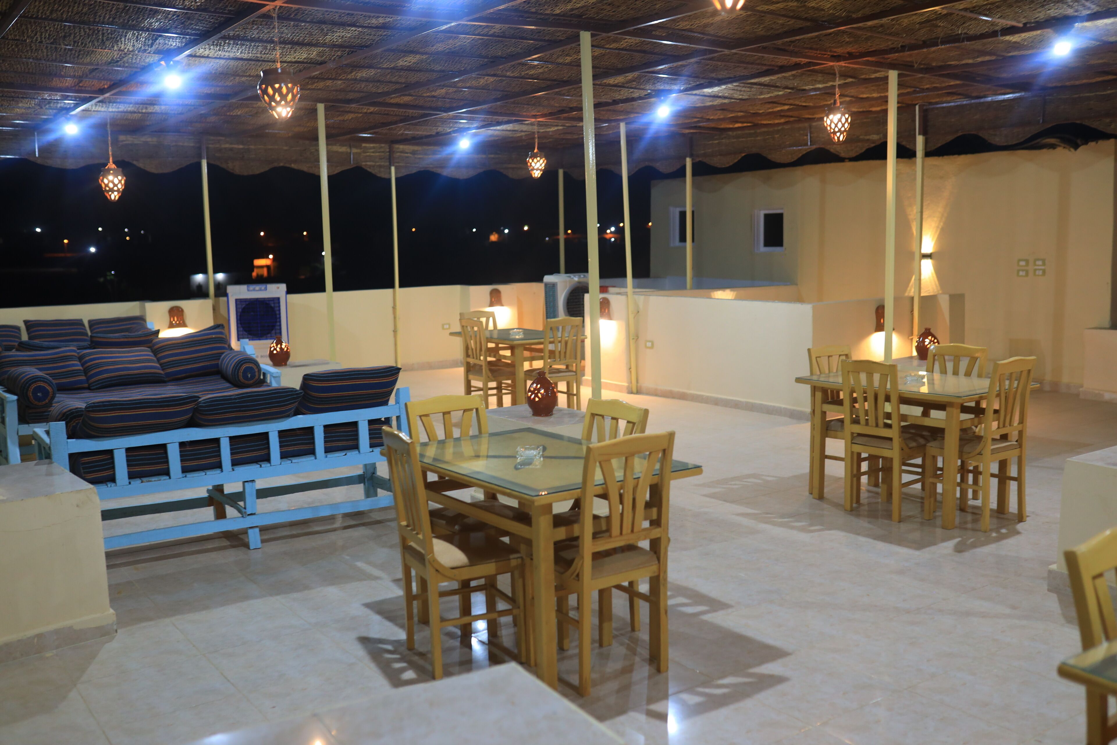 A rooftop terrace with a sofa set, dining tables, and chairs, under a thatched roof with hanging lights.