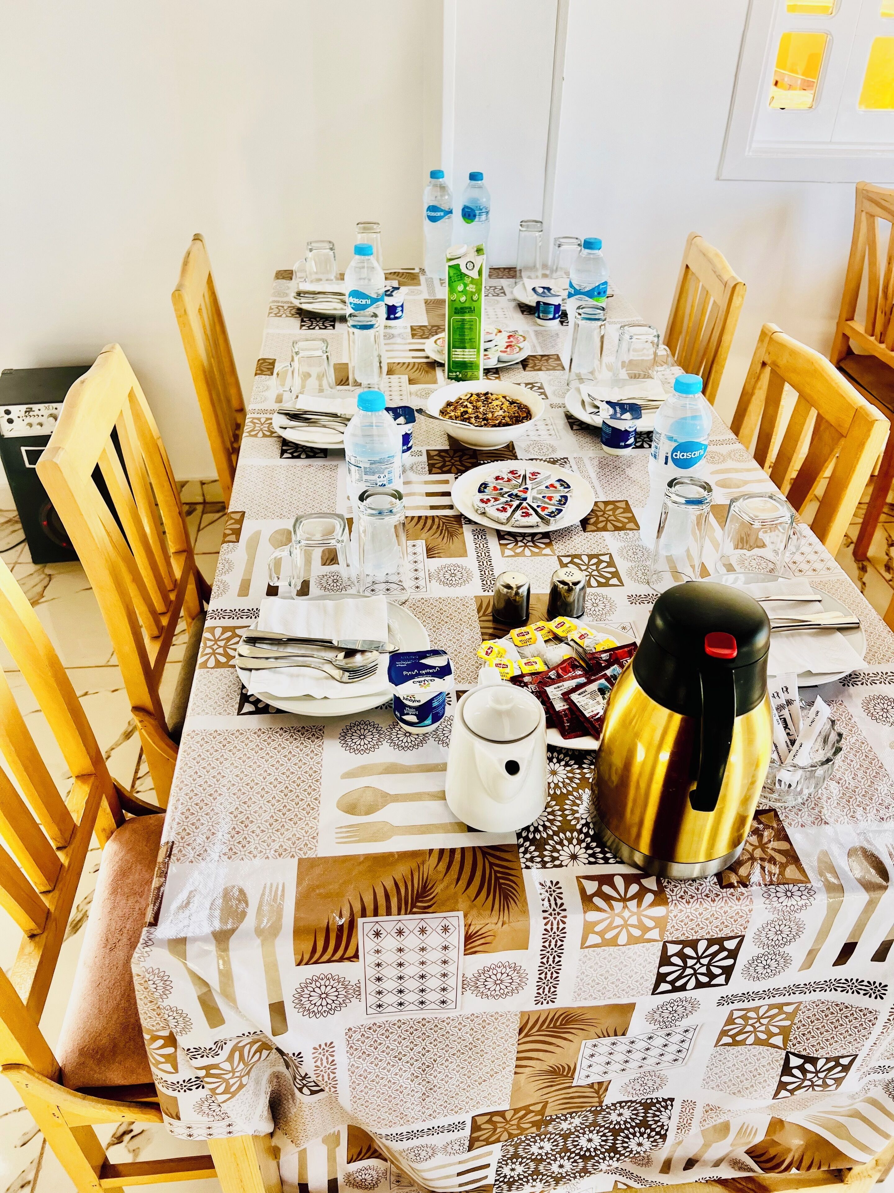 Une table à manger dressée avec des assiettes, des couverts, des bouteilles d’eau, une cafetière et un saladier rempli de nourriture.