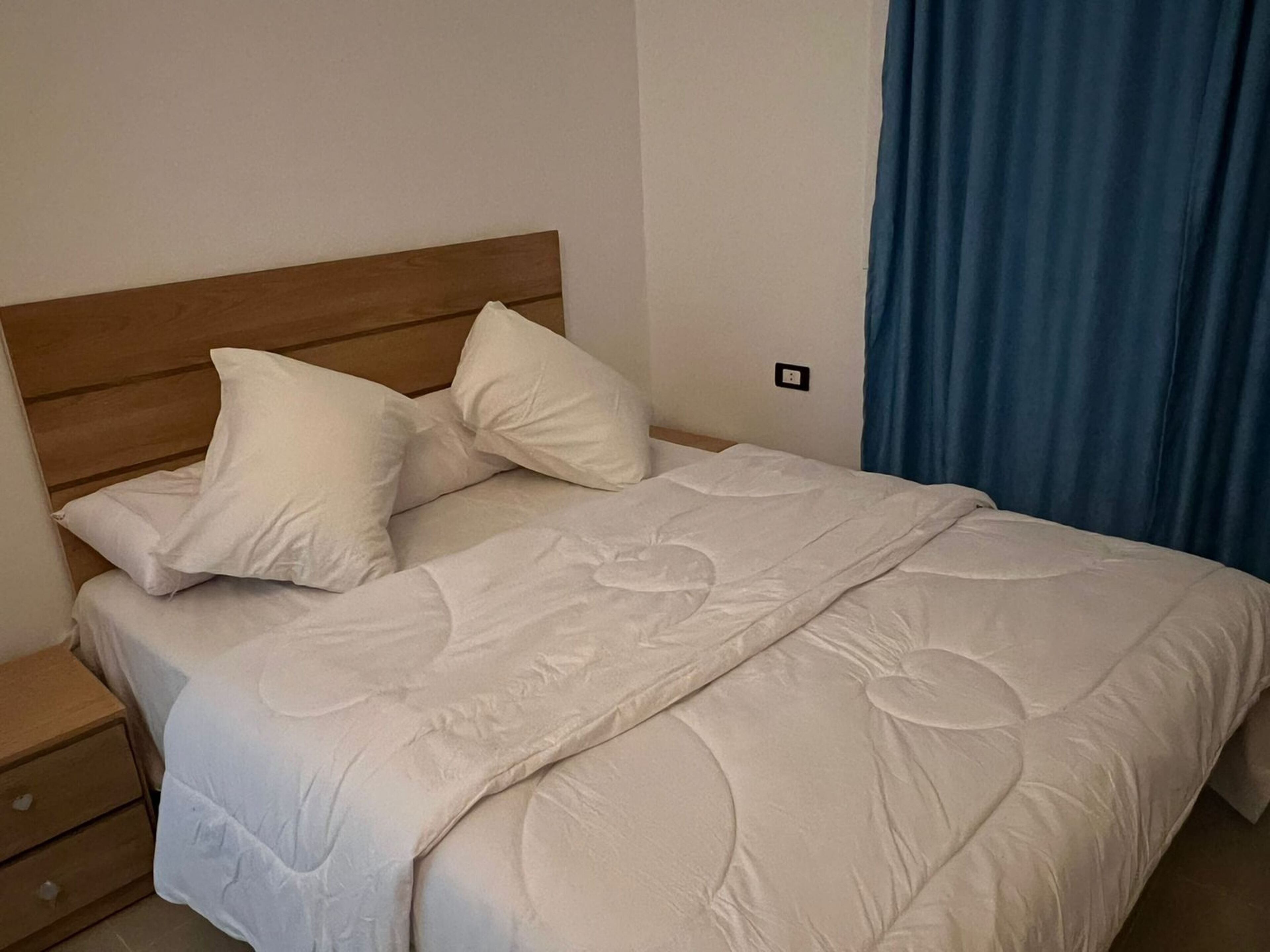 A neatly made bed with white bedding and pillows, a wooden headboard, and a blue curtain on the right.