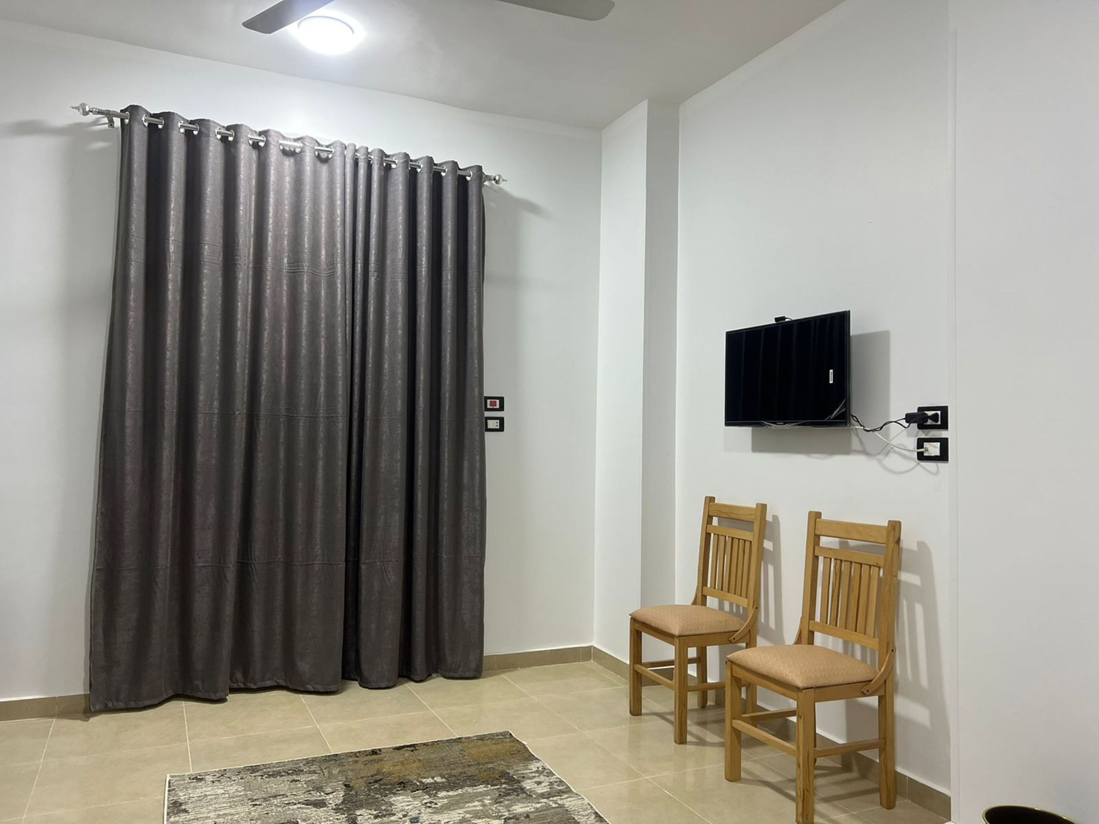 A room with a curtain, a television mounted on the wall, and two wooden chairs.