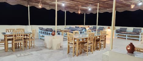 An outdoor dining area with wooden tables and chairs, a thatched roof, and hanging lights.
