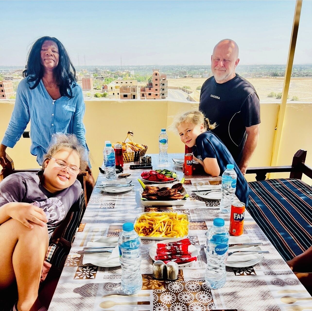 Un groupe de personnes est assis autour d’une table, avec de la nourriture et des boissons, sur un balcon donnant sur une vue panoramique de la ville.