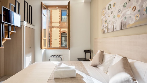 A hotel room with a large bed, a TV, a chair, and a window with a view of a brick building. - DELEDDA HOME (Cagliari)