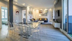 A modern living room with a glass dining table, clear chairs, a sofa, and a TV mounted on the wall. - Beautiful 120m2 Flat in bourgeois building with terrace (Cannes)