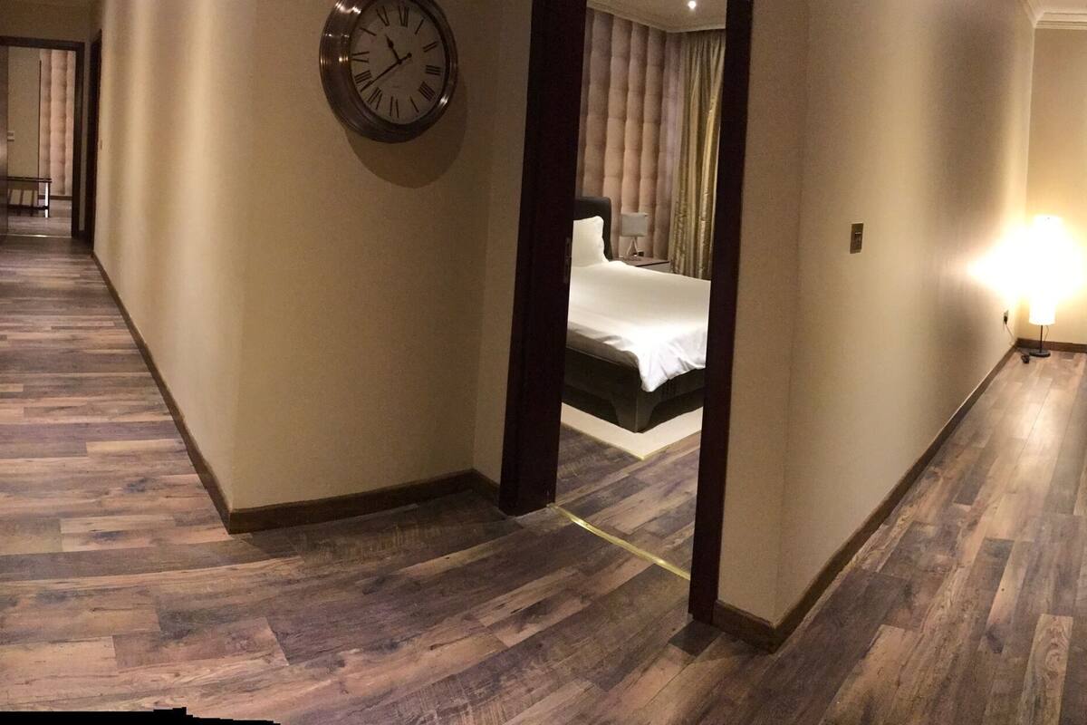A hotel hallway with a wooden floor, a bed with a headboard, a sitting area with a chair and table, and a clock on the wall.