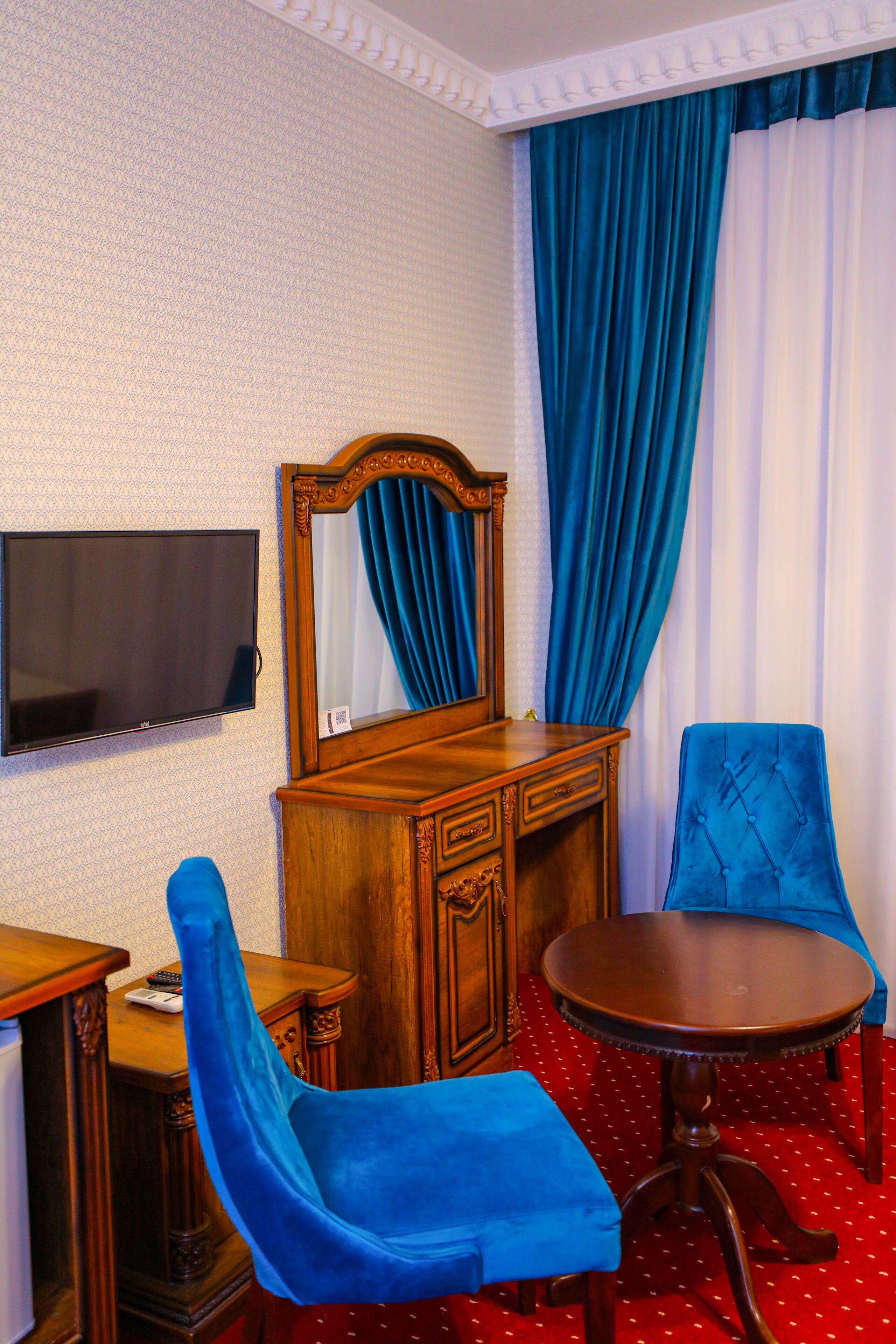 Een kamer met een houten dressoir, een aan de muur bevestigde televisie, een blauwe stoel, een ronde tafel en een groot raam met blauwe gordijnen.
