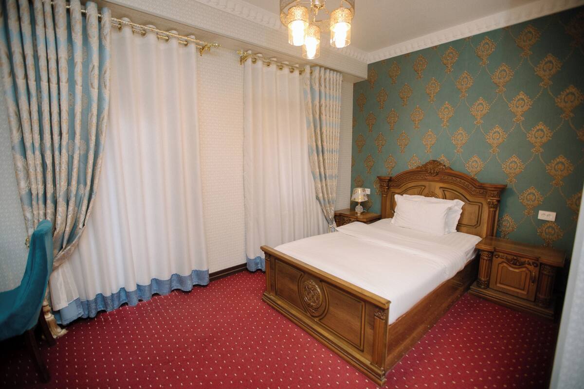 A neatly made bed with a wooden headboard, a nightstand with a lamp, and a chair. The room has a red carpet and patterned wallpaper.