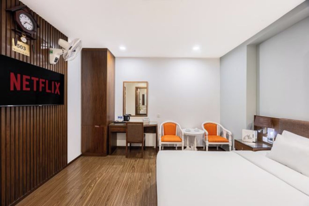 A hotel room with a bed, a desk, two chairs, a clock, and a wall-mounted TV screen displaying "NETFLIX".