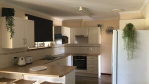 A modern kitchen with white cabinets, a central island with stools, a refrigerator with a hanging plant, and a microwave above the island. - Pelicans Rest Albany, 5 min to the beach, 6 min to city. Great all year round! (Collingwood Park)