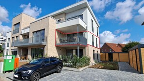 Een modern meerhoogappartementencomplex met balkons, een auto ervoor en een klein tuintje.