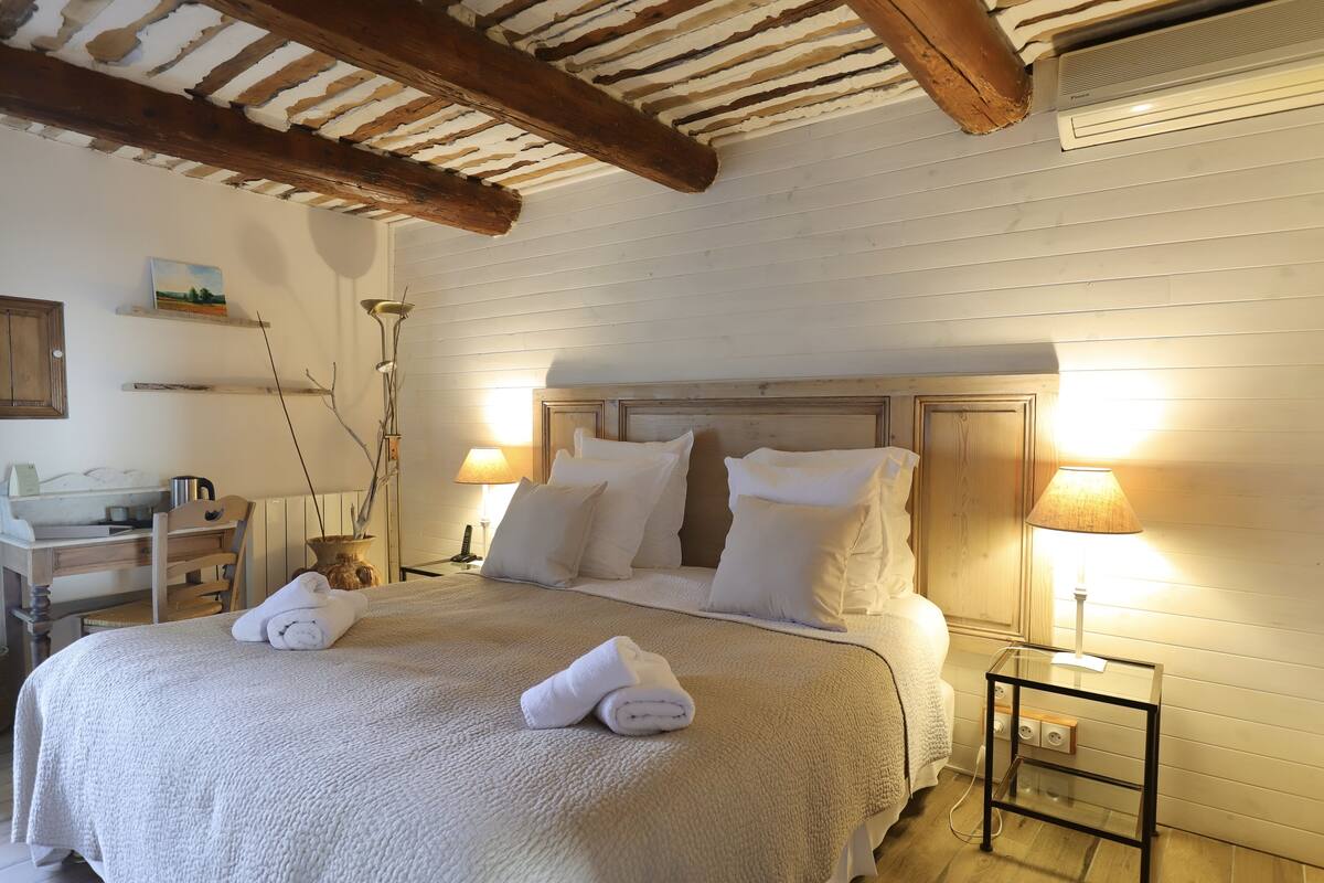 Un lit bien fait, avec du linge de lit et des serviettes blanches, un bureau en bois avec une lampe et une table de chevet avec une lampe.