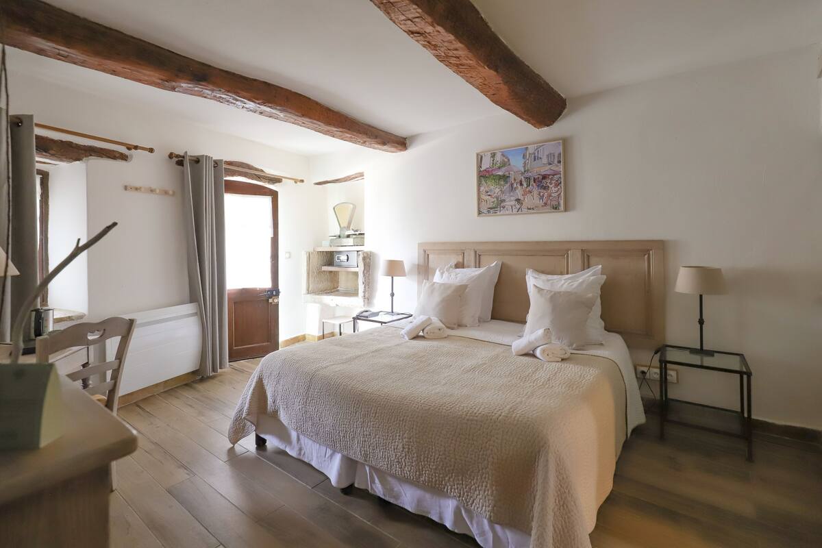 Une chambre à coucher avec un grand lit, des poutres apparentes, une table de chevet, une lampe et un tableau au mur.