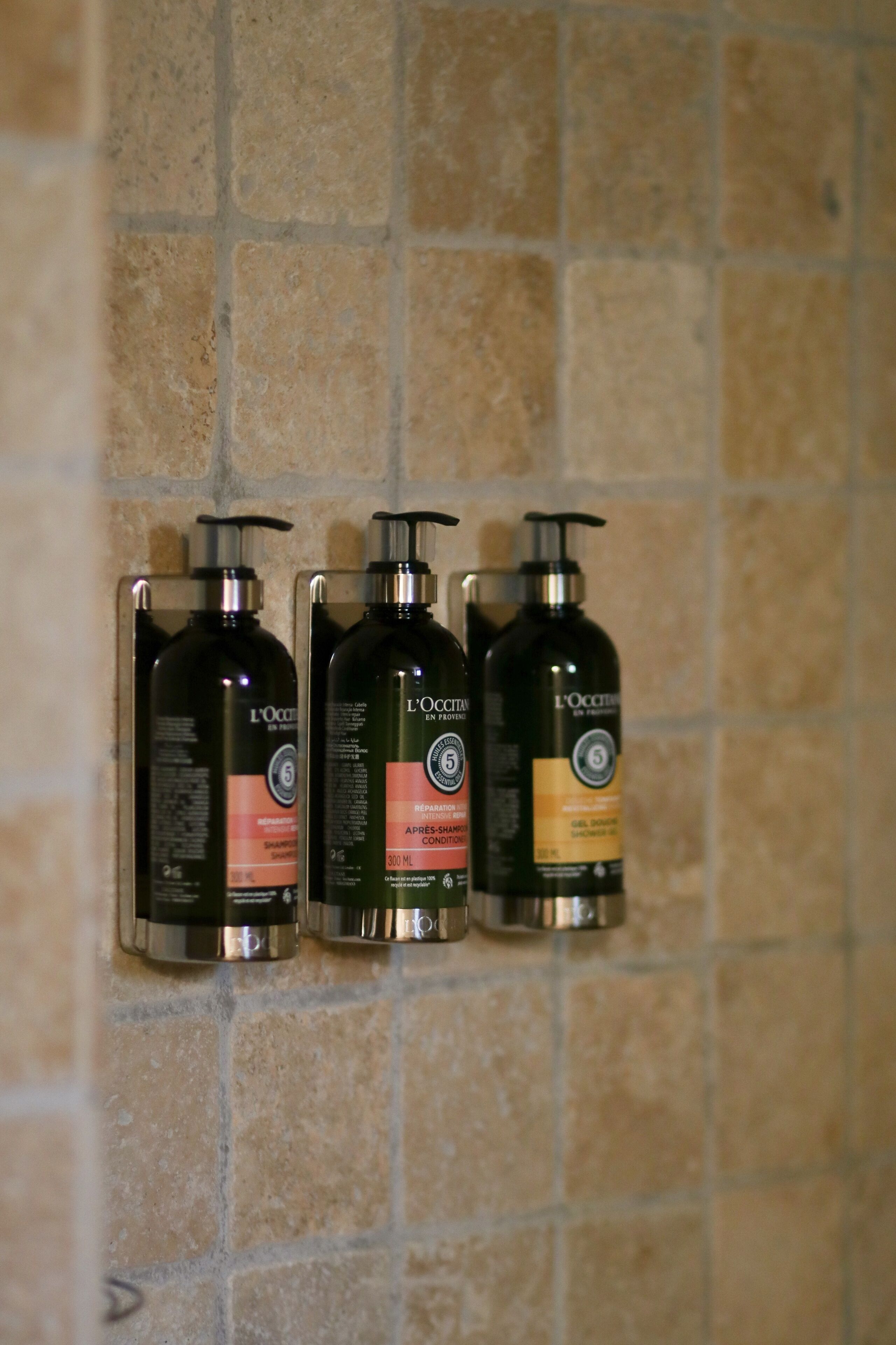 Three L'OCCITANE shower bottles mounted on a tiled wall.
