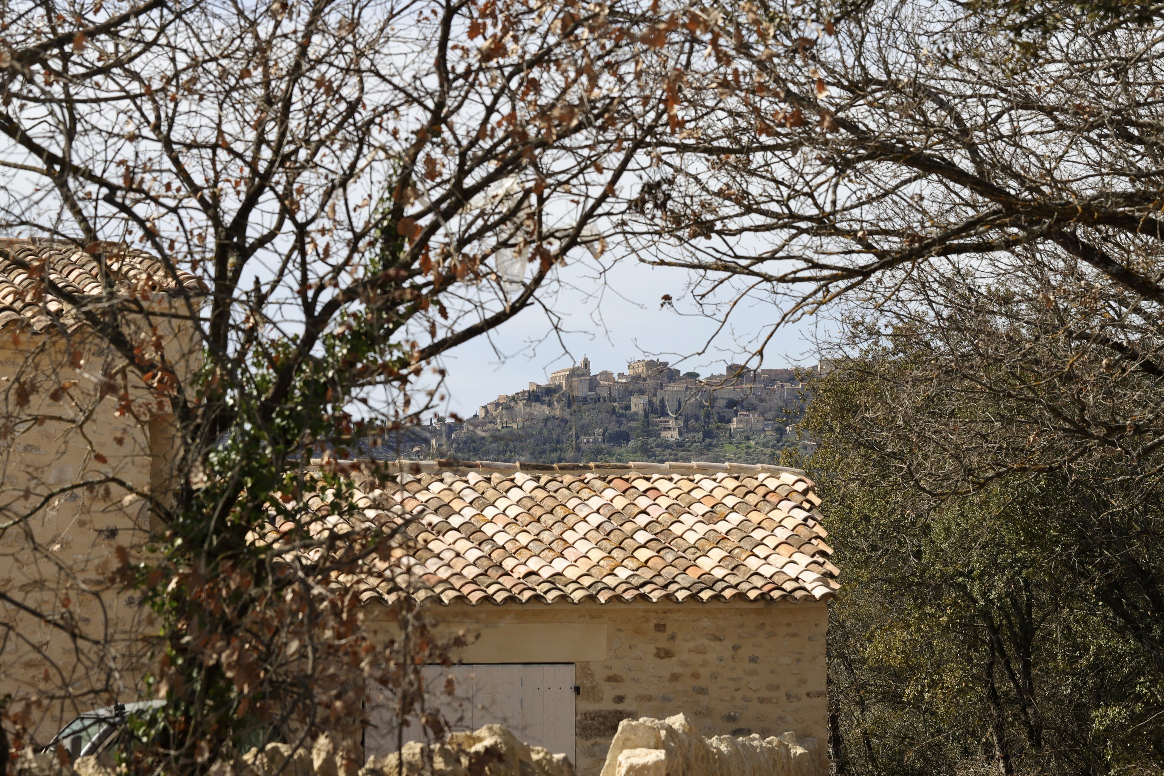 A stone building with a tiled roof, surrounded by trees and overlooking a hillside with buildings.