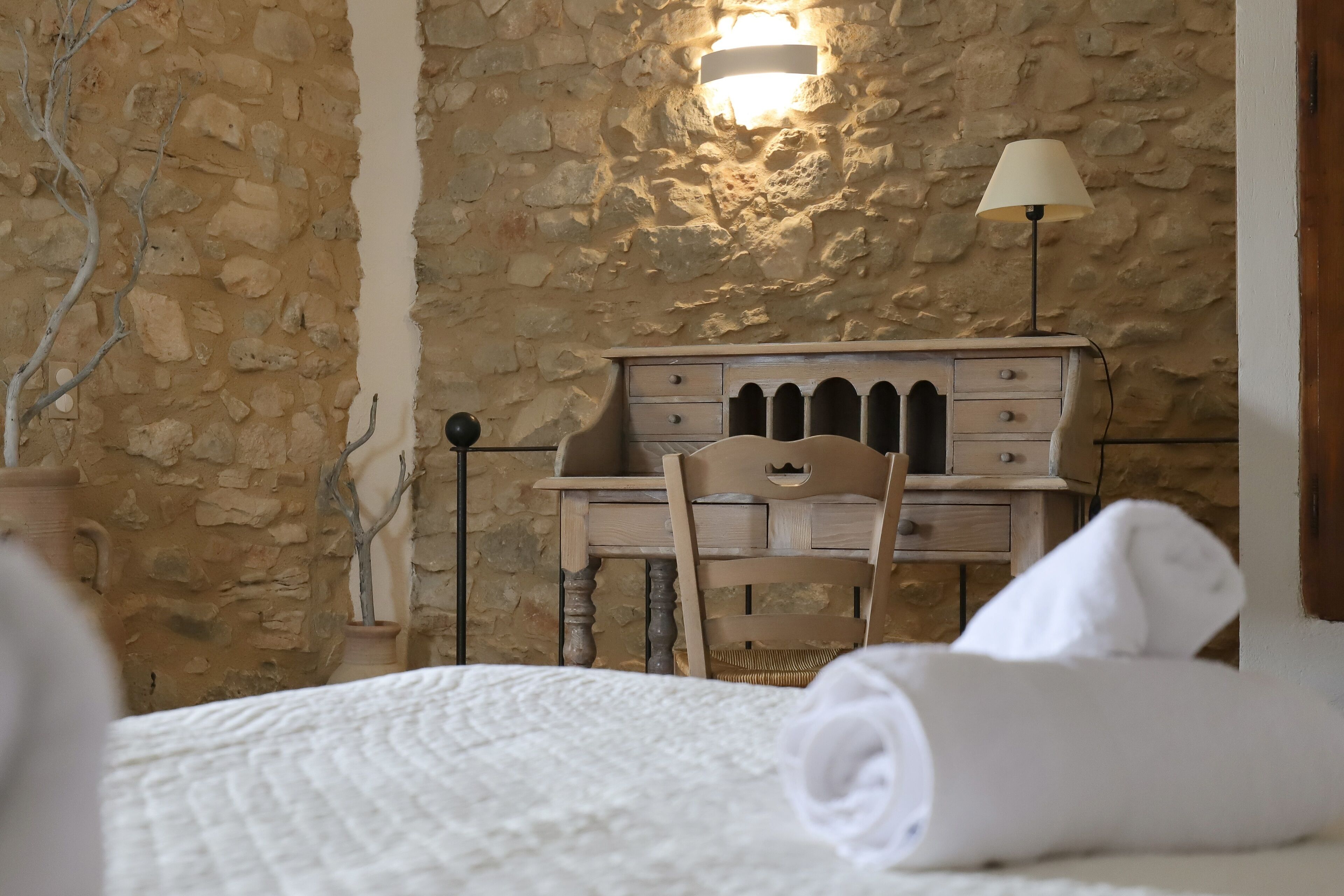 Ein Schlafzimmer mit Steinwand, einem Holztisch mit Schubladen, einem Stuhl, einer Lampe und einem zusammengerollten Handtuch auf dem Bett.