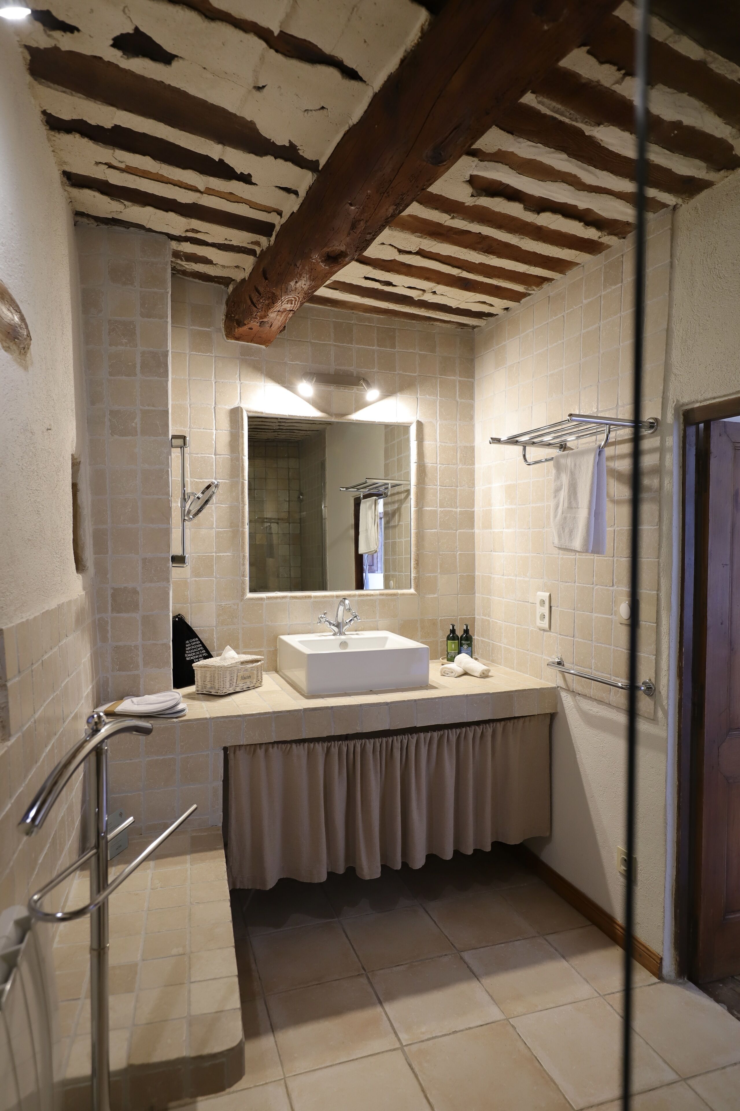 Un baño con lavamanos blanco, espejo y paredes con azulejos. Hay un perchero y una zona de ducha.