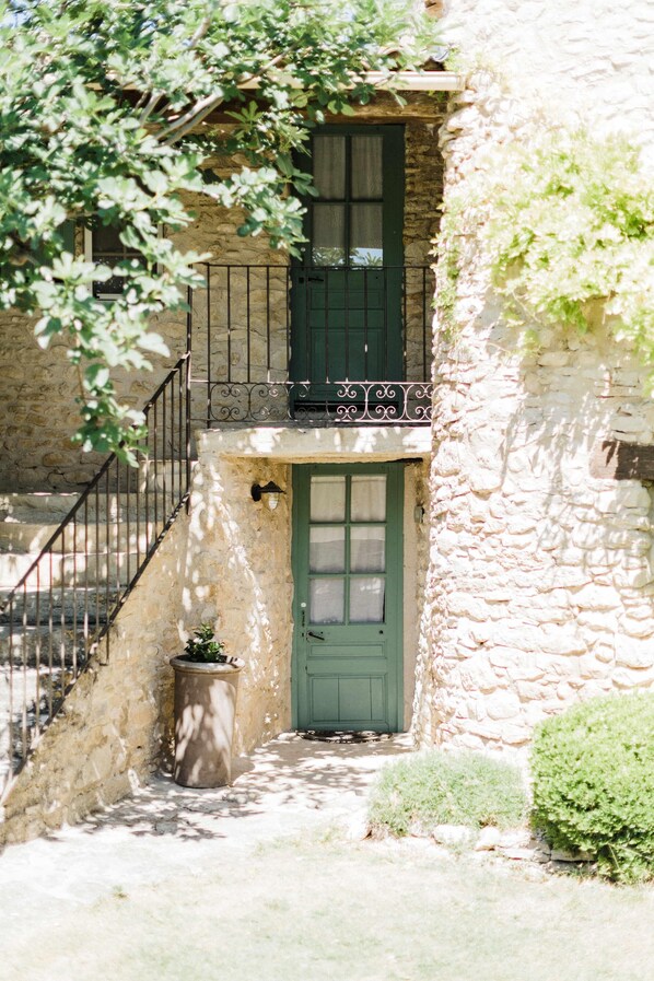Un edificio de piedra con puerta verde, un pequeño balcón con barandilla de hierro forjado y una escalera que sube a un nivel superior.
