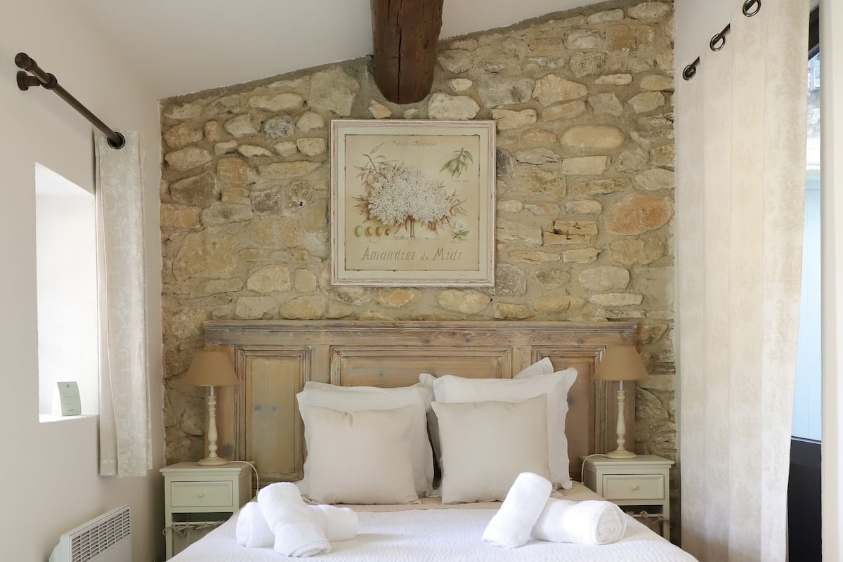 Une chambre avec un mur en pierre, une poutre en bois au plafond, un lit avec du linge de lit blanc, deux tables de chevet avec des lampes et un tableau encadré au mur.